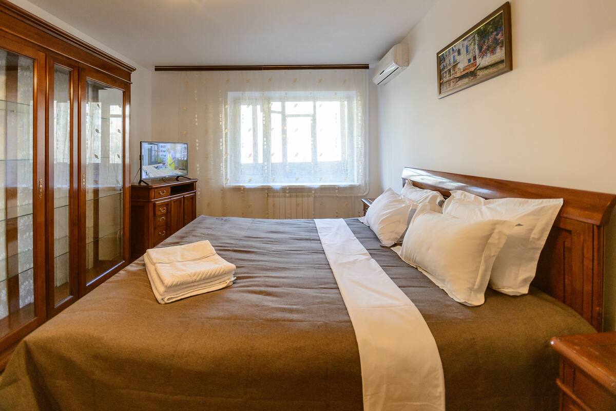 A neatly made bed with white linens, a wooden headboard, and a window with sheer curtains.