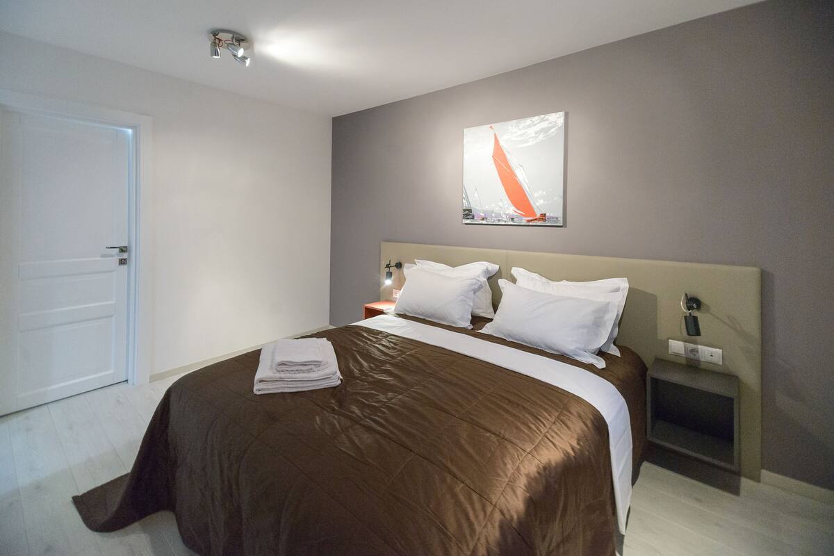 A neatly made bed with a brown bedspread and white pillows, a bedside table with a lamp, and a wall-mounted painting.