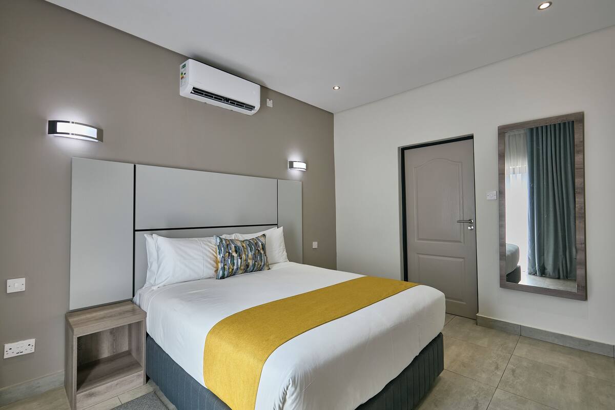 A hotel room with a large bed, a bedside table, an air conditioning unit, a door, and a mirror with curtains.