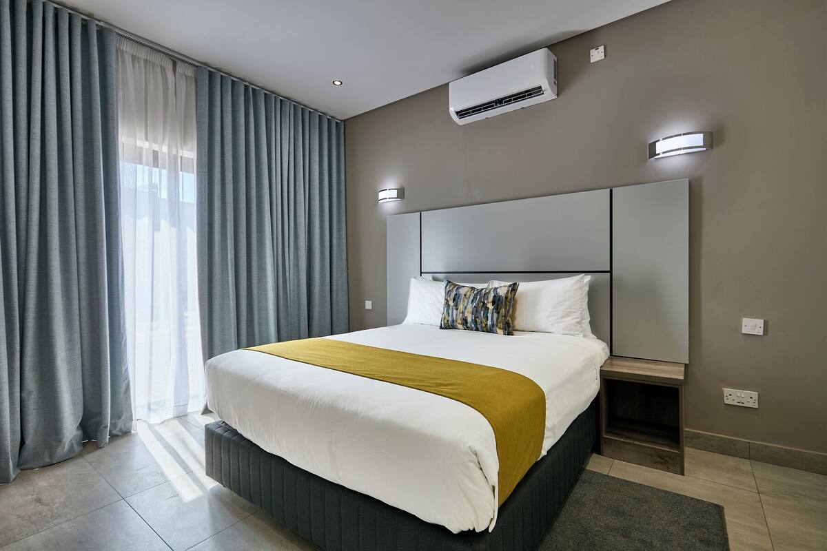 A modern hotel room with a large bed, a bedside table, an air conditioning unit, and a wall-mounted light fixture.