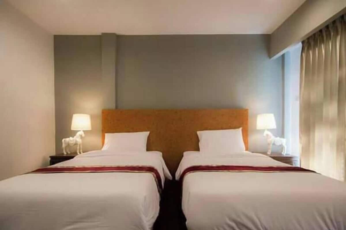 A hotel room with two single beds, a wooden headboard, bedside lamps, and a window with curtains.