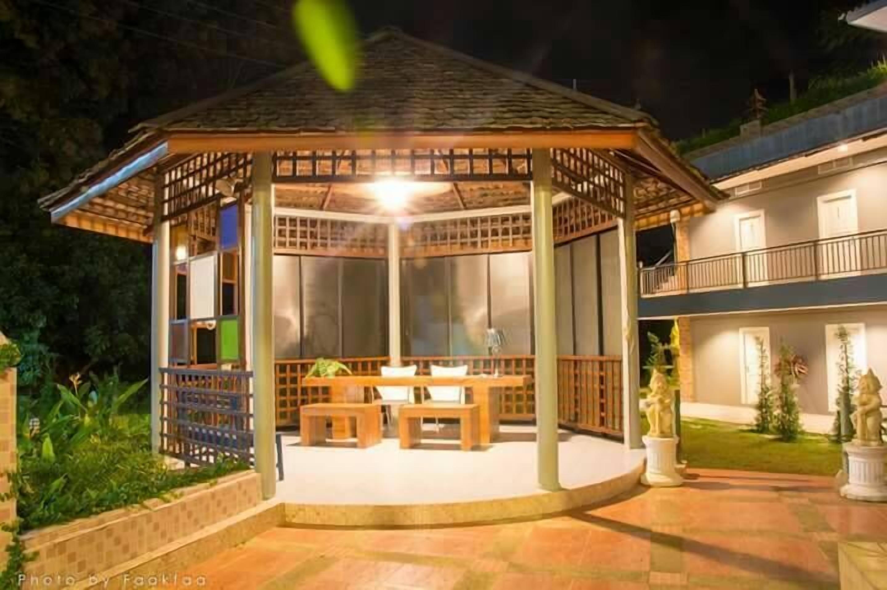 A gazebo with a tiled floor and wooden furniture, surrounded by a garden and a building with a balcony.