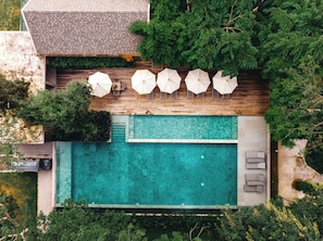 An aerial view of a swimming pool with a wooden deck, surrounded by greenery and umbrellas.