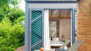 A wooden deck with two lounge chairs and a small table, a sliding glass door leading to a room with a bed and a nightstand, and a view of lush greenery. - Flora Creek Resort & Garden Chiang Mai (Chiang Mai)