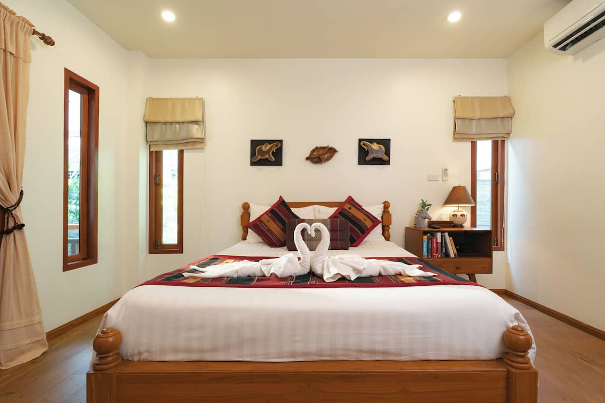 A neatly made bed with a heart-shaped towel arrangement, a bedside table with books and a lamp, and a window with a view of greenery.