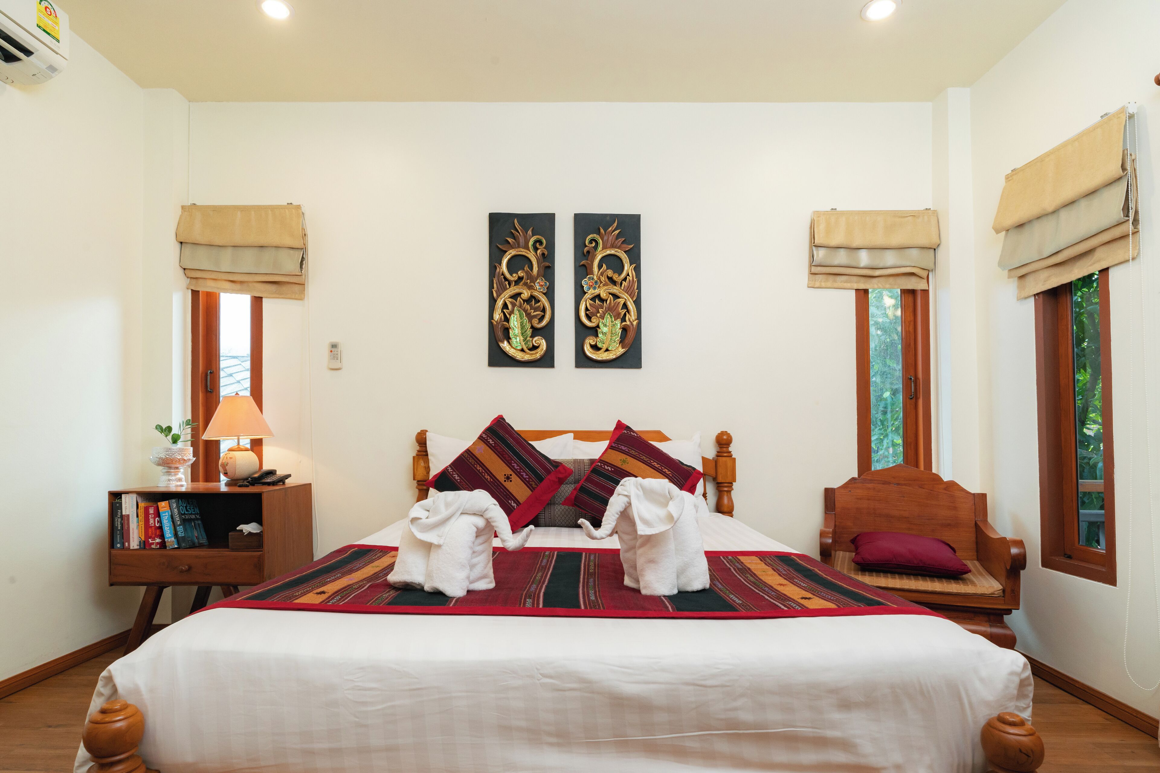 A neatly arranged bedroom with a large bed, two wall-mounted artworks, a bedside table with books, a chair, and a window with a view.