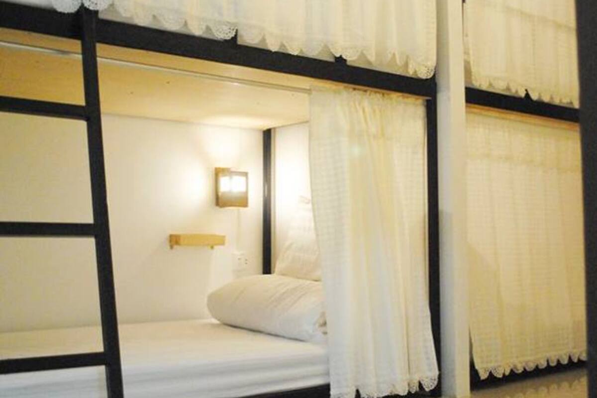Habitación con literas, ropa de cama blanca, una lámpara en la pared y cortinas translúcidas.