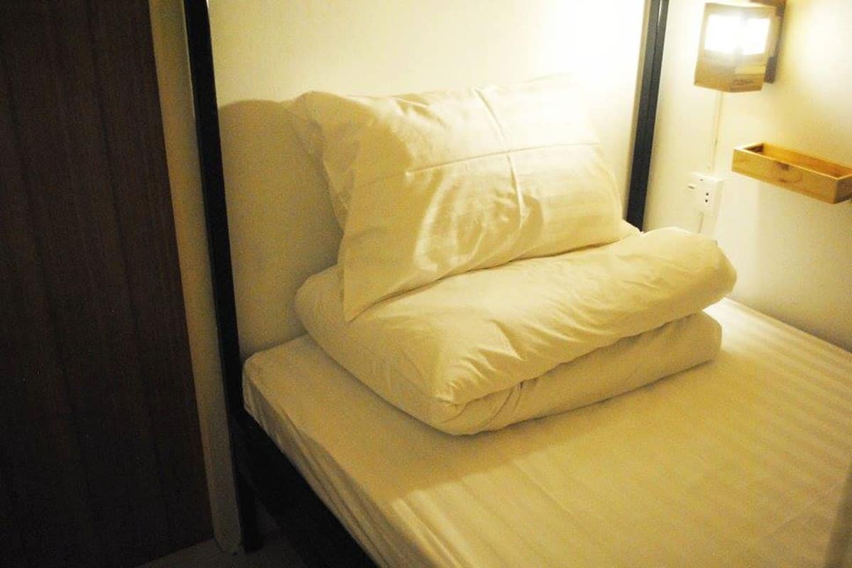 Una cama individual con ropa blanca, en una habitación con cabecera de madera y mesita de noche con lámpara.