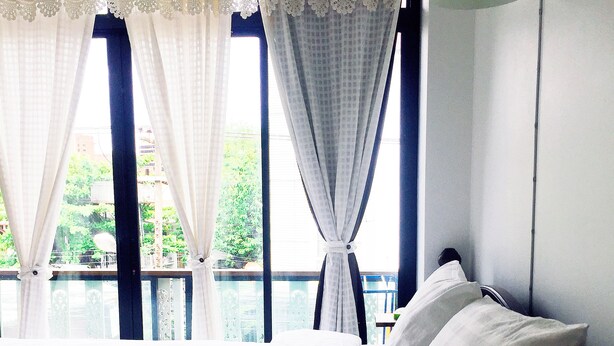 A bedroom with a bed, white curtains, a window with a view of greenery, and a wall-mounted lamp.