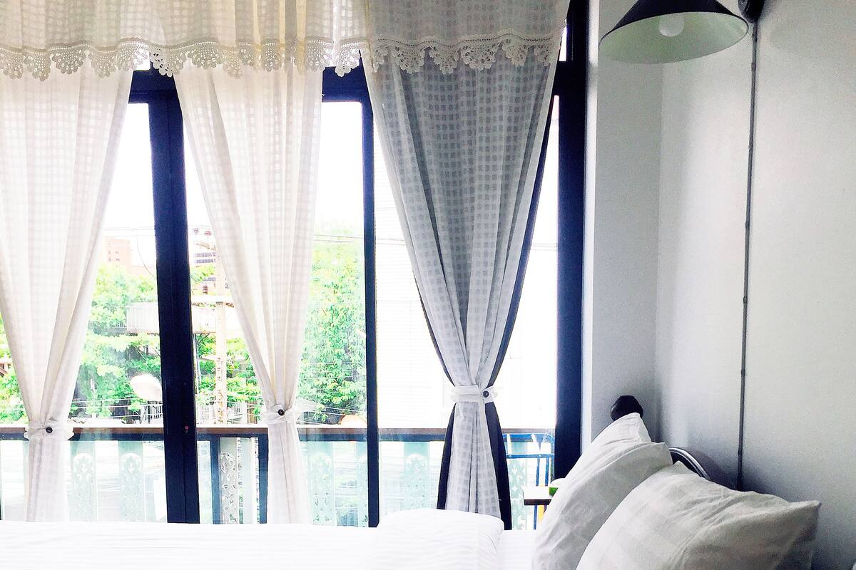 Un dormitorio con cama, cortinas blancas, una ventana con vista a follaje y una lámpara fijada a la pared.