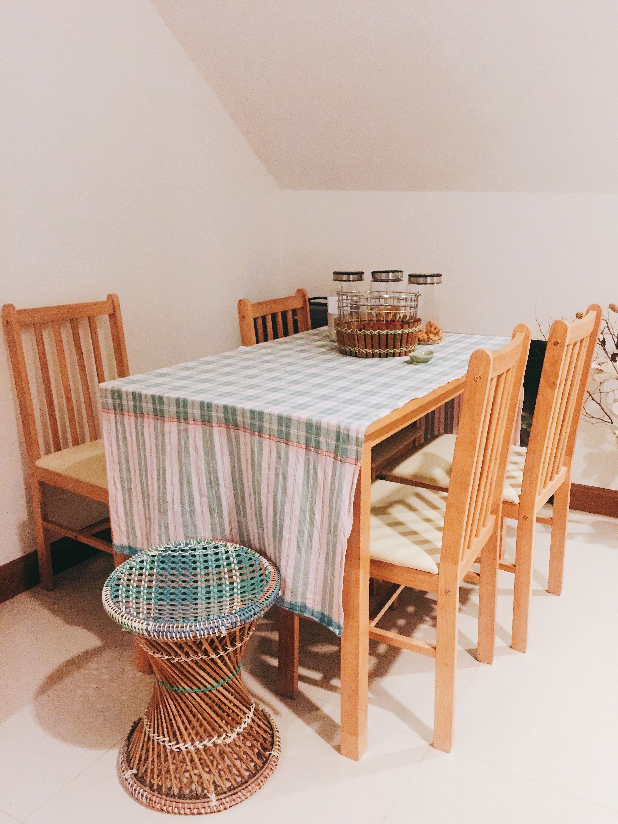 Un comedor con mantel a cuadros, sillas de madera, un taburete de mimbre y un cesto de frascos sobre la mesa.