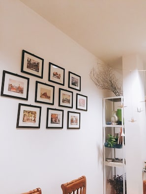 A room with a white wall displaying framed pictures, a wooden chair, and a white shelving unit with decorative items.