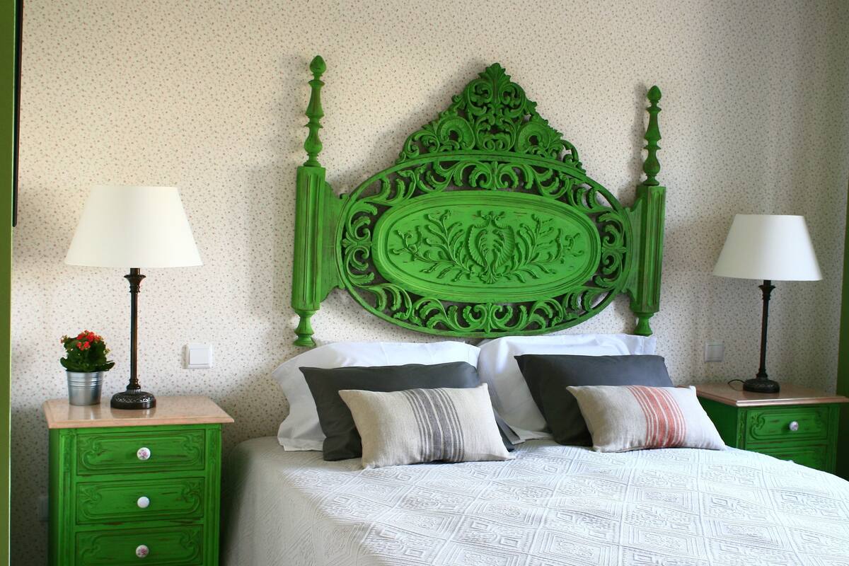 A bedroom with a green headboard, two bedside tables with lamps, a bed with patterned bedding, and a small potted plant.