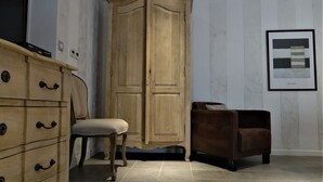 A room with a wooden wardrobe, a chair, a brown sofa, and a framed artwork on the wall.