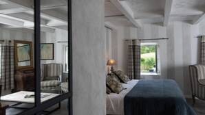 A bedroom with a bed, a chair, a table, and a window with a view of greenery.
