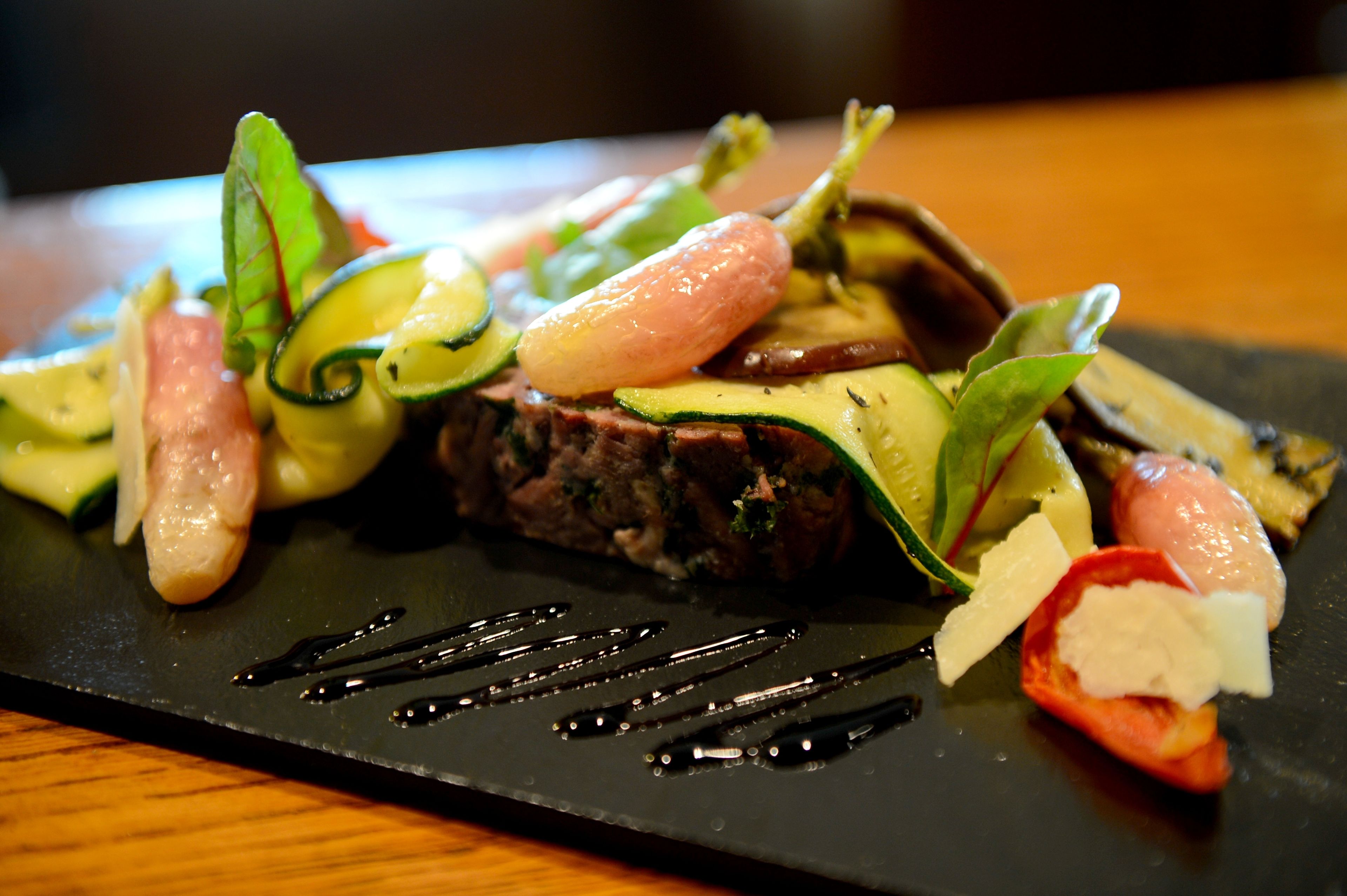 A black slate plate with a variety of vegetables and a piece of meat, possibly steak, garnished with herbs and a slice of tomato.