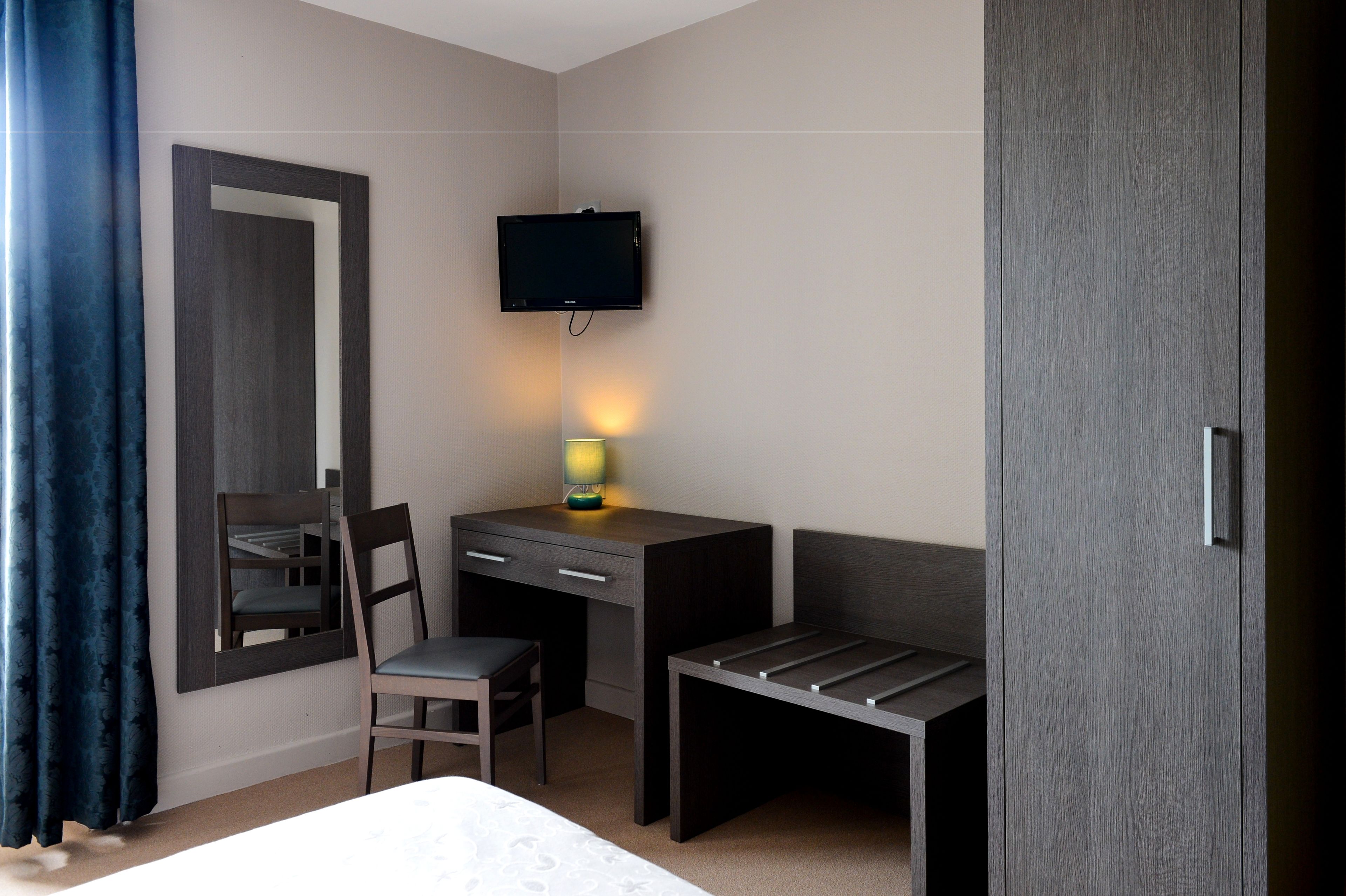 A hotel room with a bed, a desk with a chair, a TV mounted on the wall, and a wardrobe.
