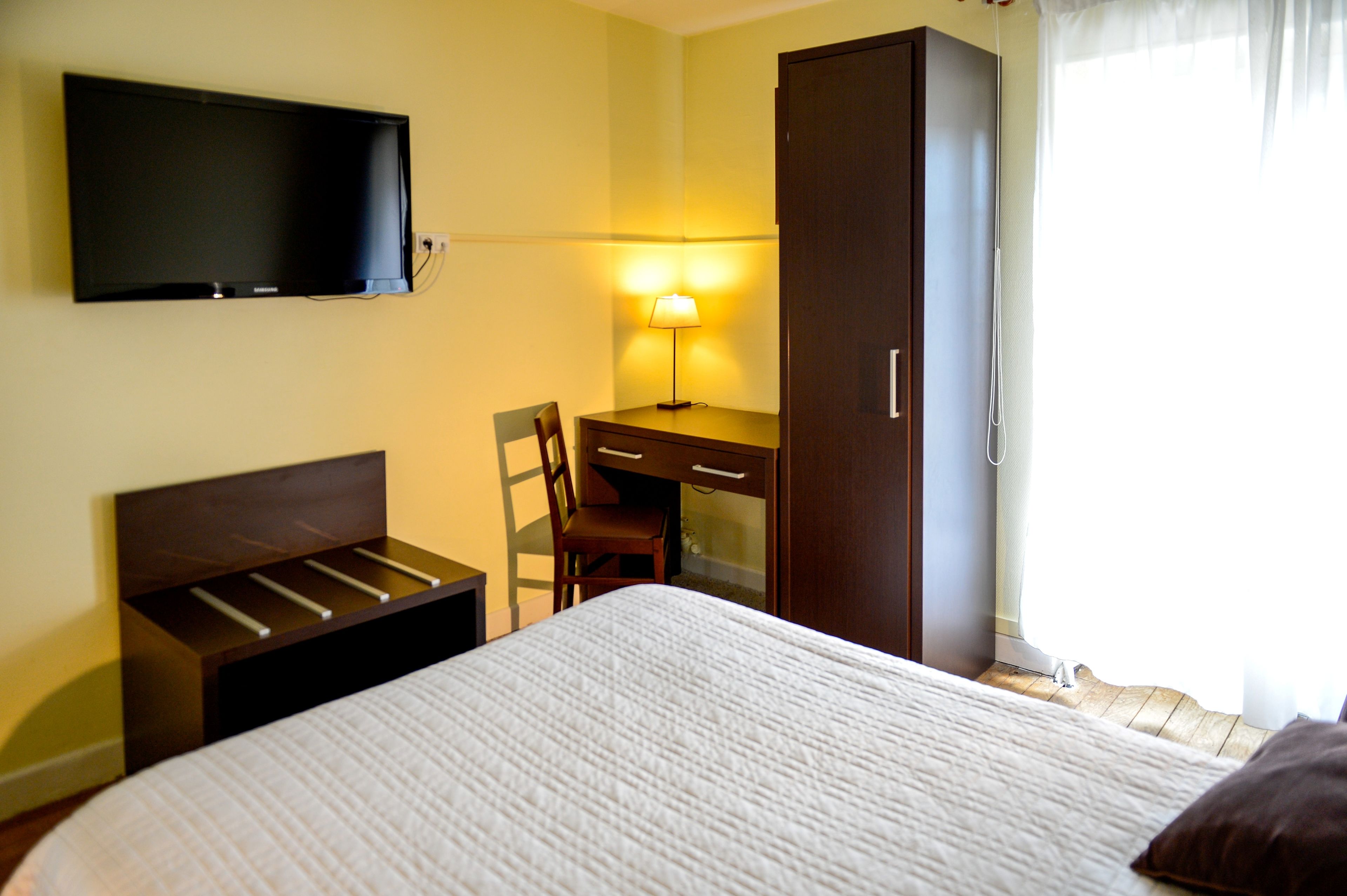 A hotel room with a bed, a desk with a chair, a television mounted on the wall, and a wardrobe.