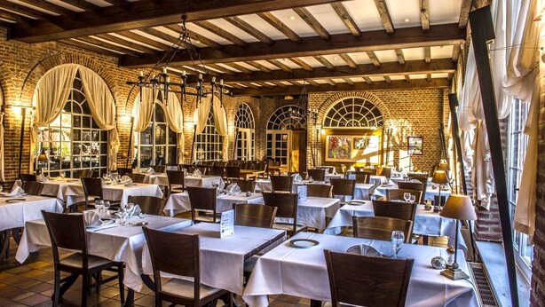 A restaurant with multiple tables, wooden beams, brick walls, and large windows with curtains. - Logis hôtel du château de la motte (Liessies)