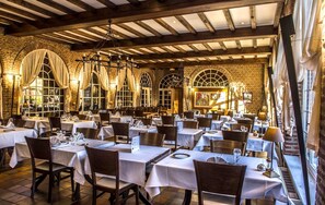 A restaurant with multiple tables, wooden beams, brick walls, and large windows with curtains. - Logis hôtel du château de la motte (Liessies)