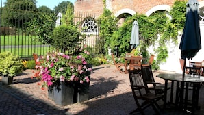 An outdoor patio area with brick flooring, wooden chairs, and tables. There is a metal gate with a decorative arch, and a brick building with arched windows in the background. - Logis hôtel du château de la motte (Liessies)
