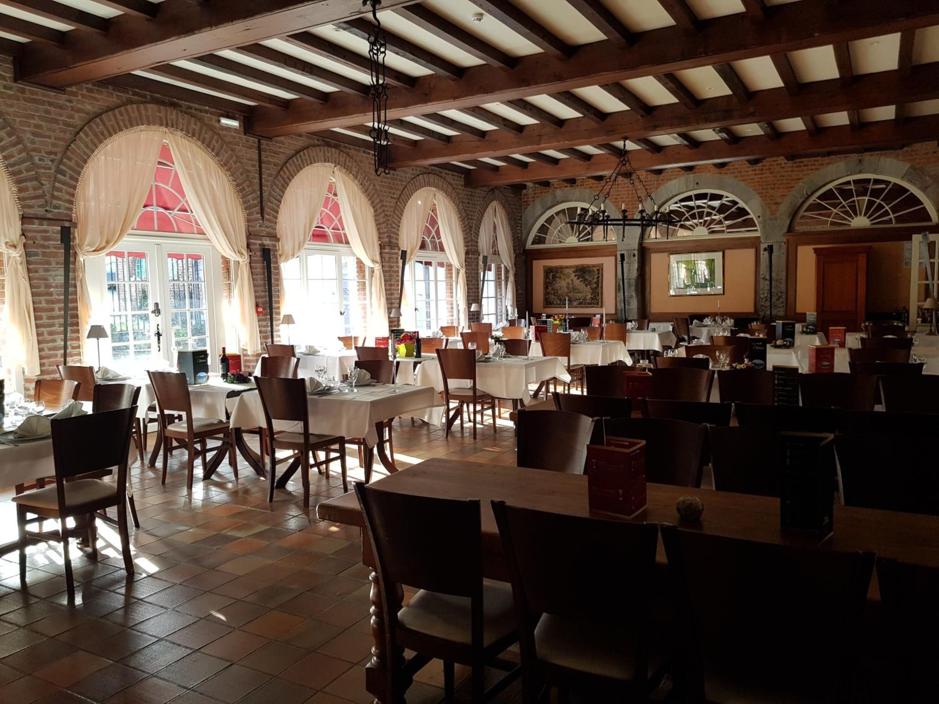 A spacious restaurant with high ceilings, exposed beams, and brick walls. Tables are set with chairs, and large windows with curtains allow natural light.