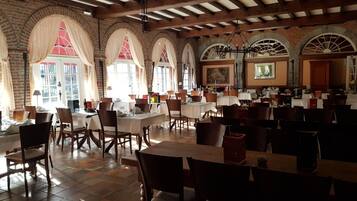A spacious restaurant with high ceilings, exposed beams, and brick walls. Tables are set with chairs, and large windows with curtains allow natural light.