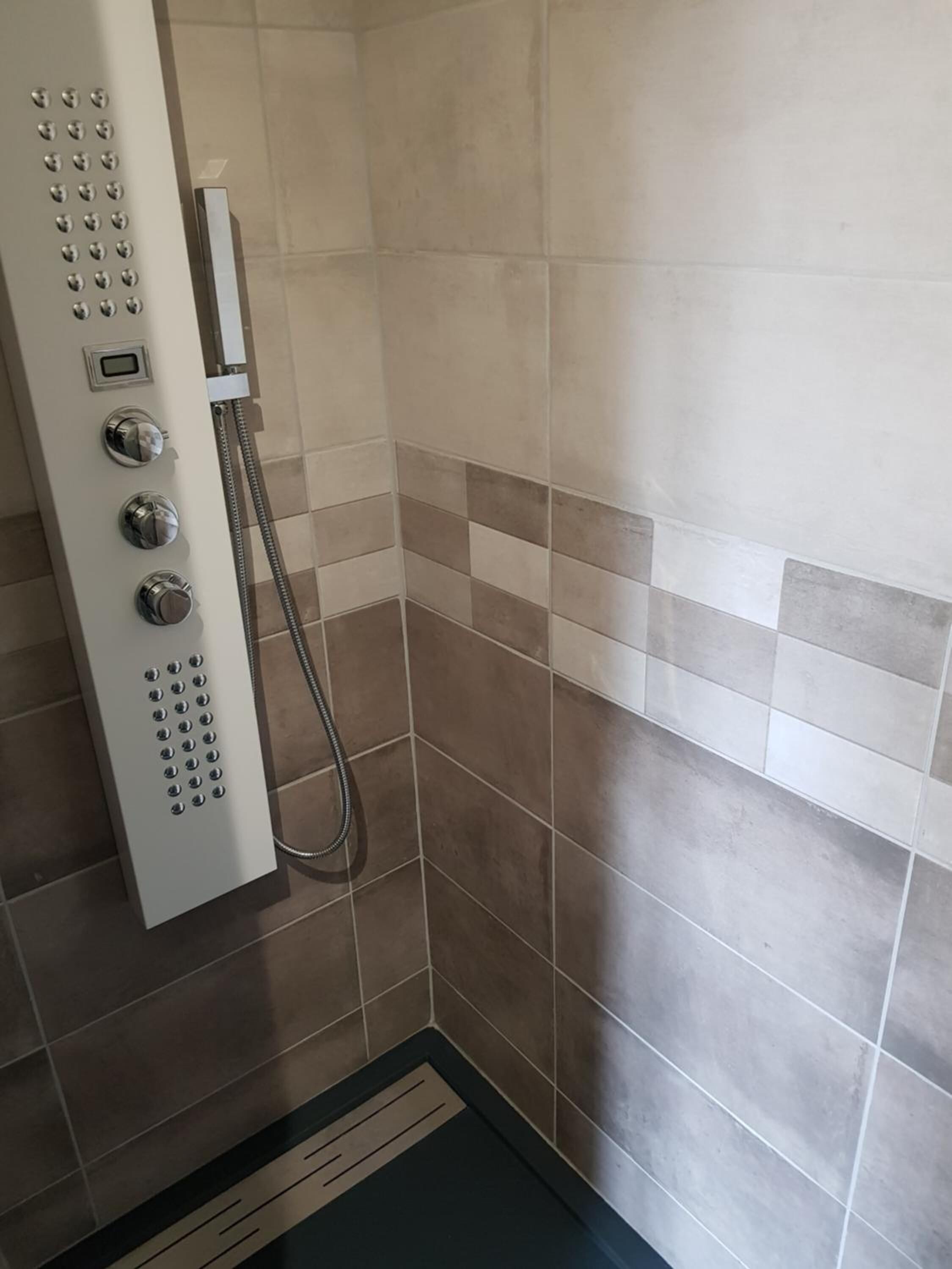 A shower with a control panel, tiled walls, and a drain.