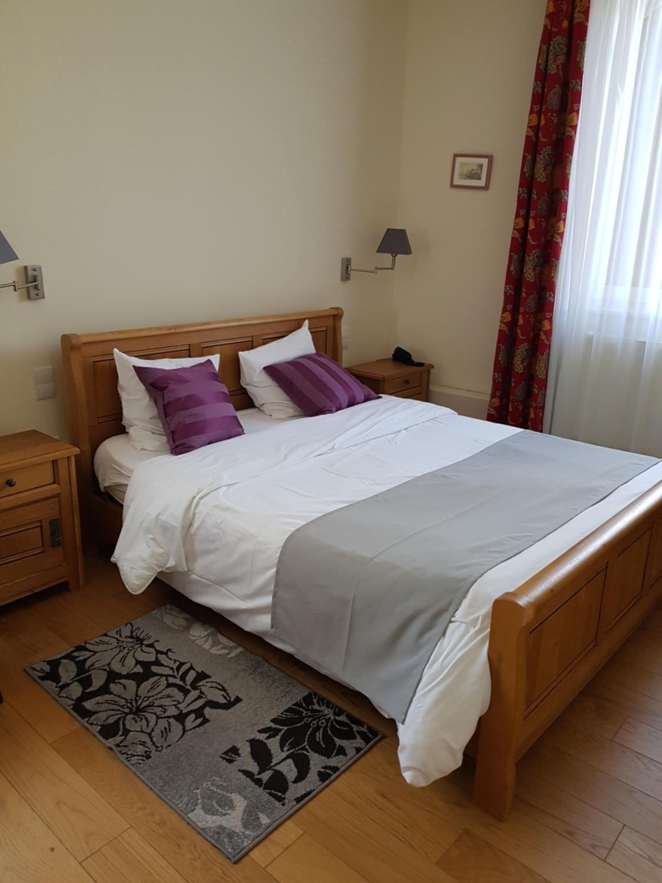 A neatly made bed with a wooden headboard, a nightstand with a lamp, and a floral rug on a wooden floor.