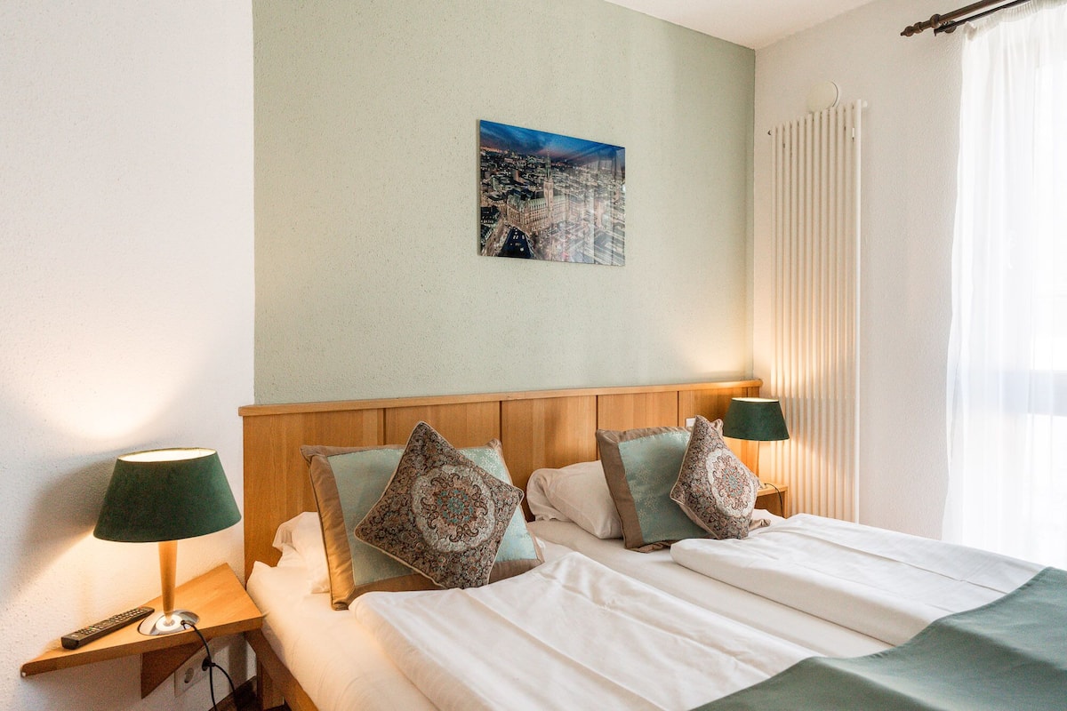 Una camera d'hotel con una testiera in legno, due comodini con lampade, un letto con lenzuola bianche e un quadro raffigurante una vista cittadina.