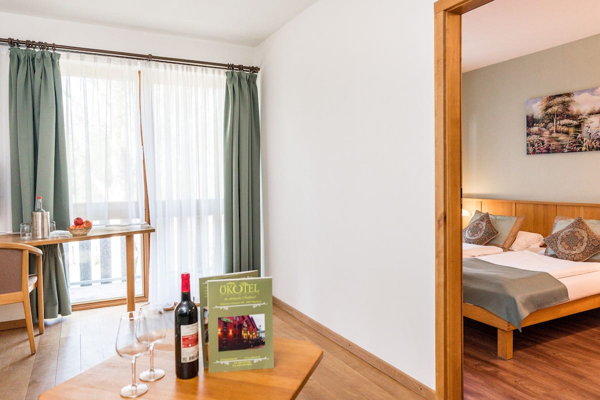 Camera d'albergo con un letto, un tavolo con bottiglia di vino e bicchieri, una sedia, un tavolino con un cesto di frutta e una finestra con tende.