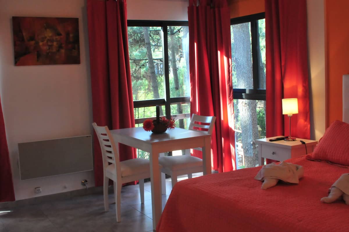 A hotel room with a bed, a desk with two chairs, a table with a vase of flowers, and a window with red curtains.