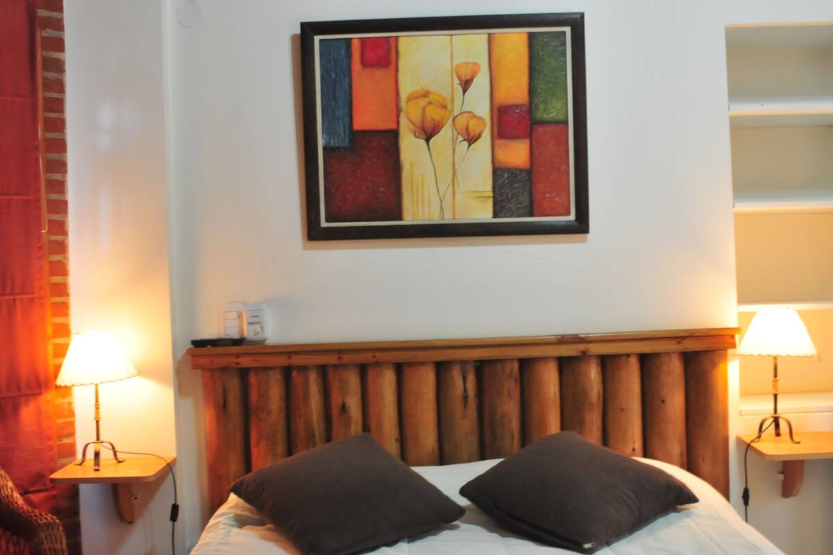 Habitación de hotel con cabecera de madera, dos lámparas de noche, un cuadro en la pared y una cama con dos almohadas.
