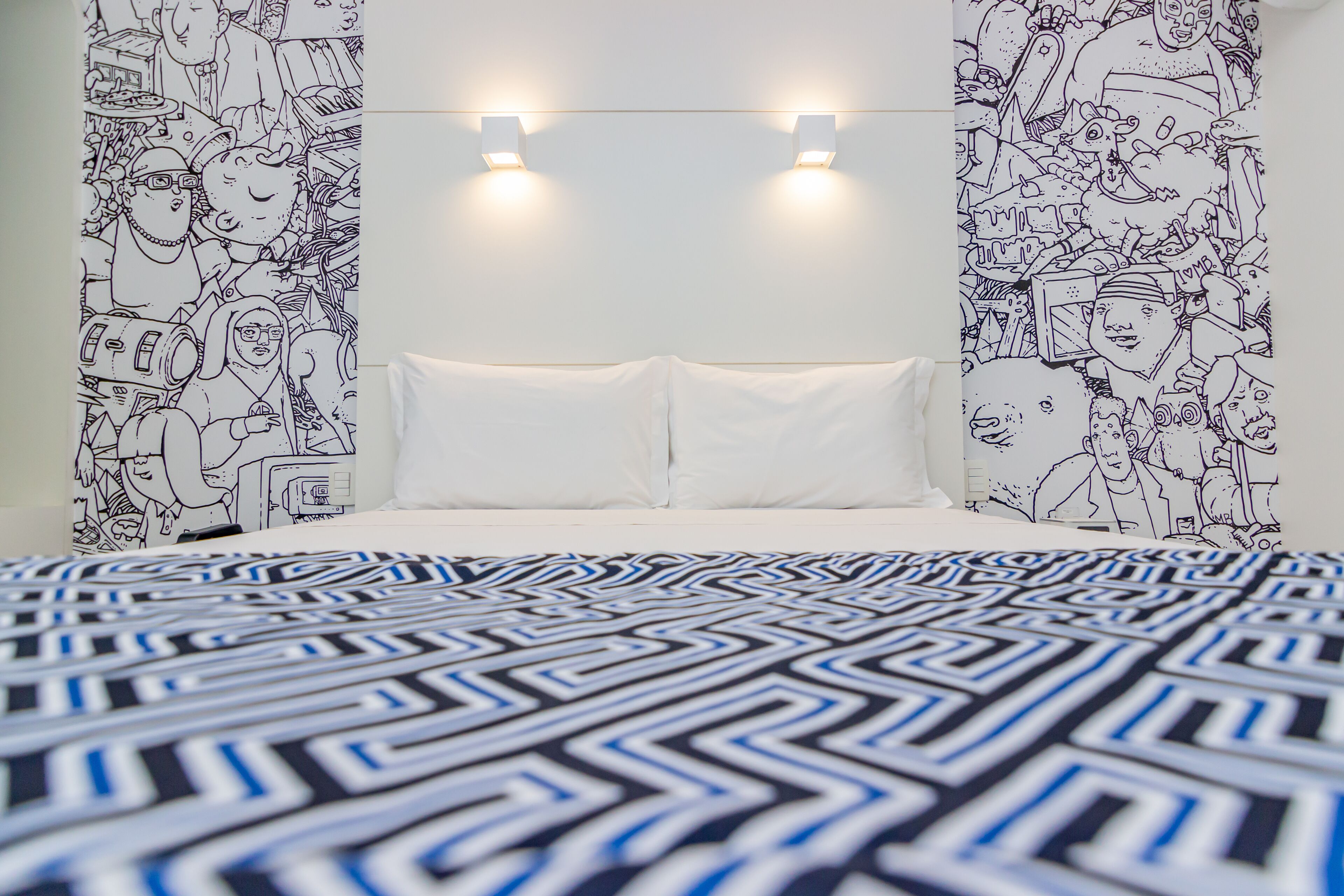 A bedroom with a patterned bedspread, white pillows, and walls decorated with black and white illustrations.