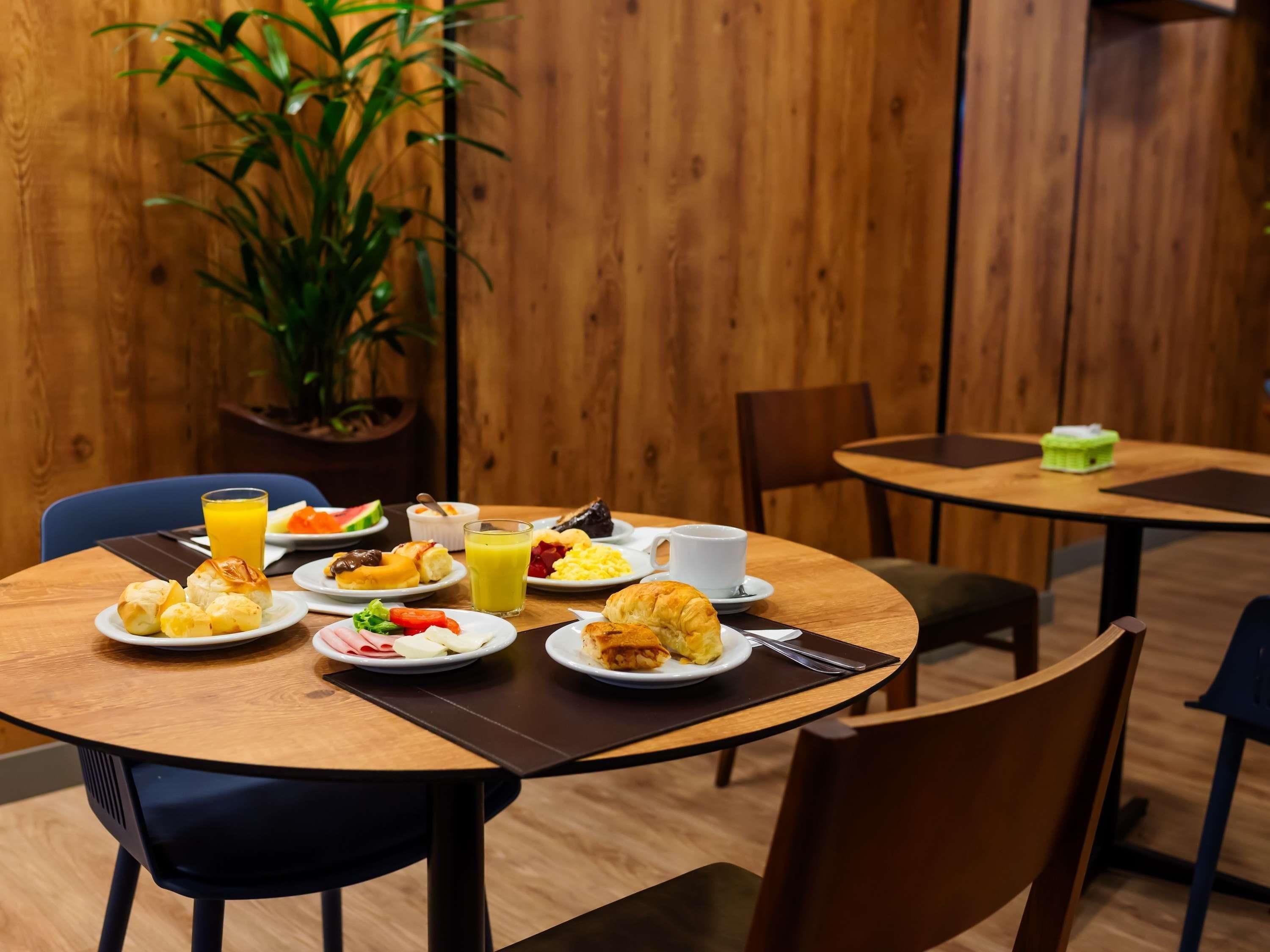 Un área de comedor con mesas y sillas de madera, una planta en maceta y diferentes opciones de desayuno, como cruasanes, huevos y jugo.