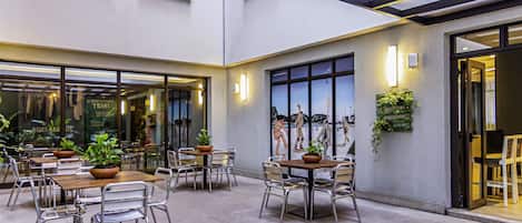 An outdoor cafe area with tables and chairs, potted plants, and a mural on the wall.