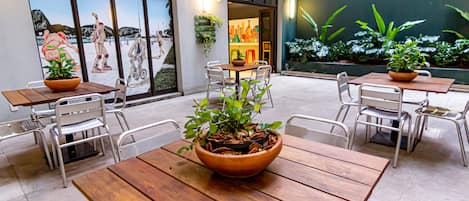 An outdoor patio area with wooden tables and chairs, potted plants, and a view of a beach through large windows.