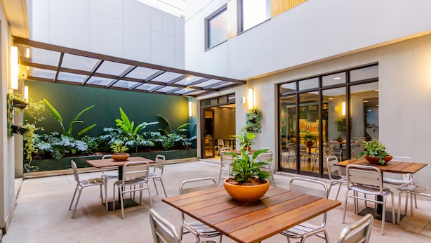An outdoor patio area with wooden tables and metal chairs, surrounded by potted plants and a glass enclosure. - Ibis Styles RJ Botafogo (Rio de Janeiro)