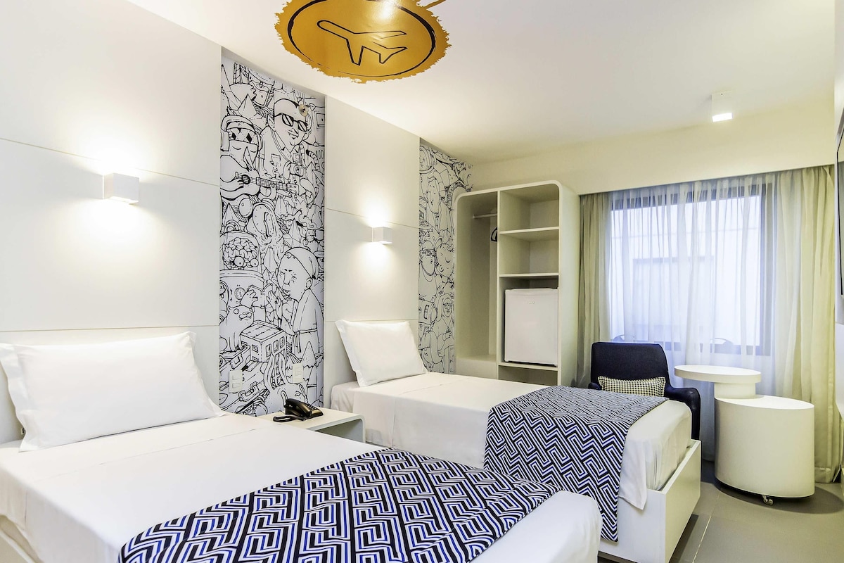 A hotel room with two beds, a desk, a chair, a wardrobe, and a window with curtains.