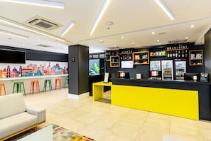 A modern bar area with a yellow counter, a cityscape mural, and a seating area with a sofa and stools. - Ibis Styles RJ Botafogo (Rio de Janeiro)