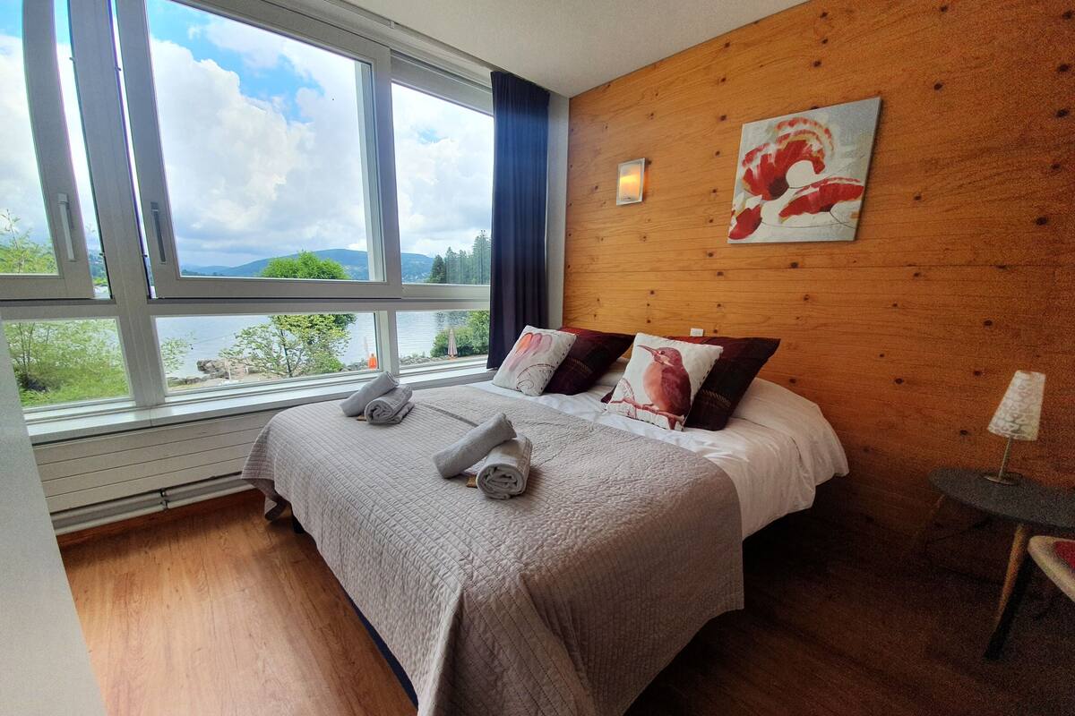 A bedroom with a large window, wooden walls, a bed with pillows, and a lamp.