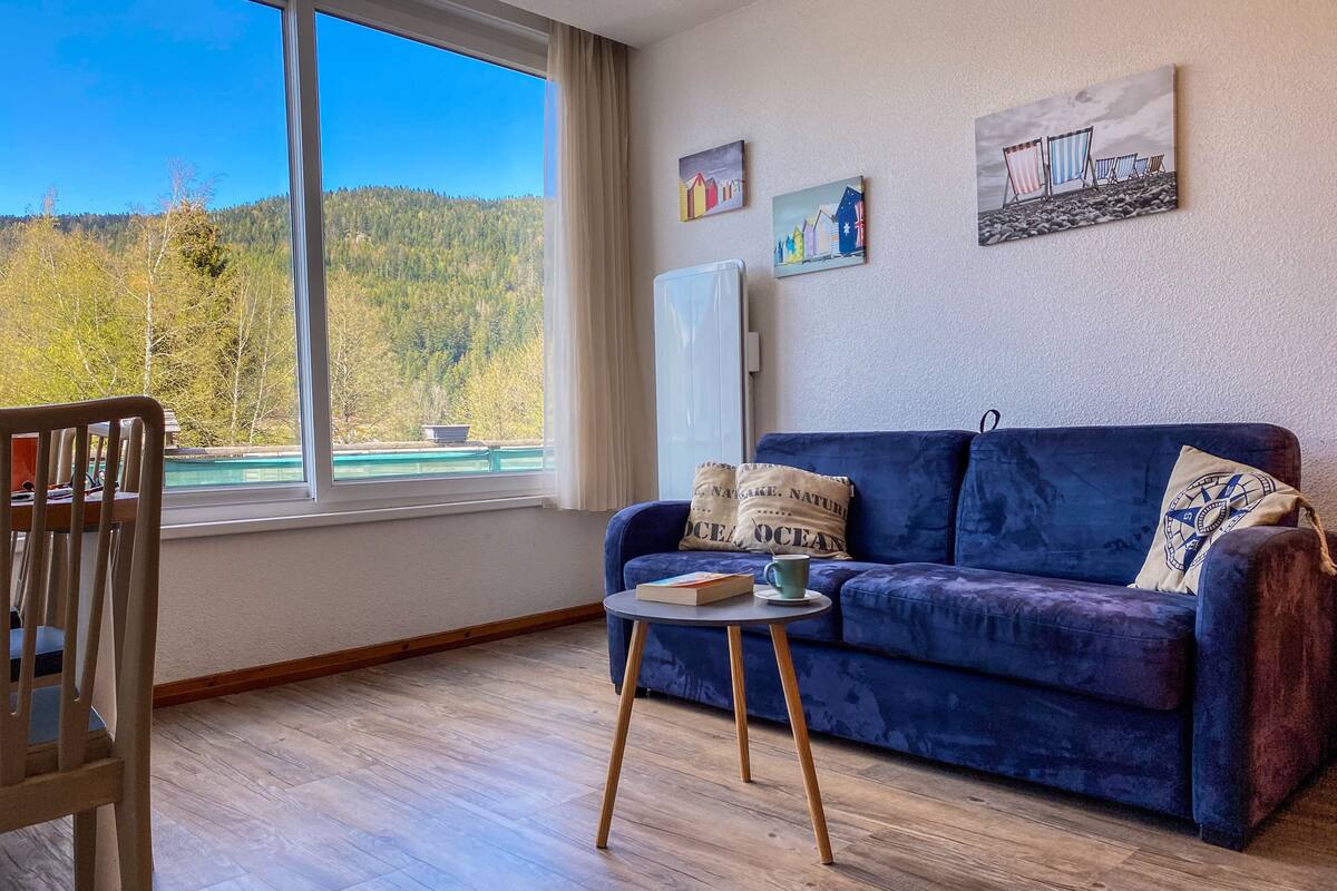 A room with a blue sofa, a small table, and a view of a forest through a large window.
