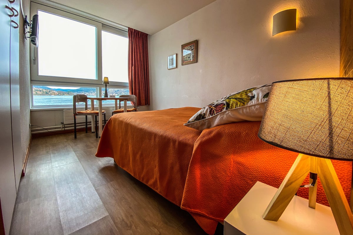 A hotel room with a bed, a desk, a chair, a lamp, and a view of the water.