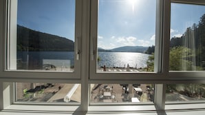 Vista a través de una ventana de una zona junto al lago con mesas y sillas, una playa de arena y una colina boscosa al fondo.
