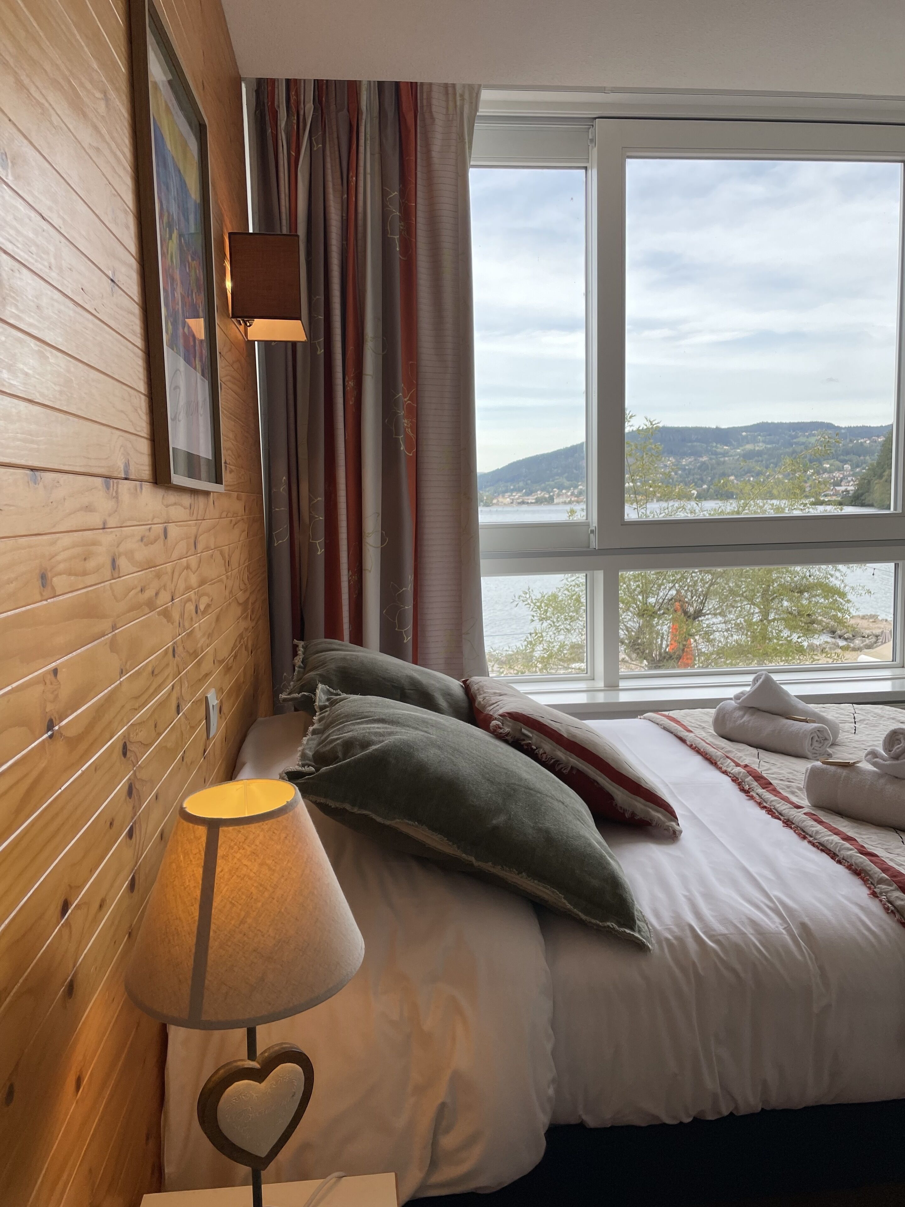 A bedroom with a wooden wall, a bed with white bedding, a heart-shaped lamp, and a view of a landscape through a window.