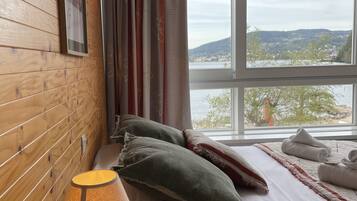 A bedroom with a wooden wall, a bed with white bedding, a heart-shaped lamp, and a view of a landscape through a window.