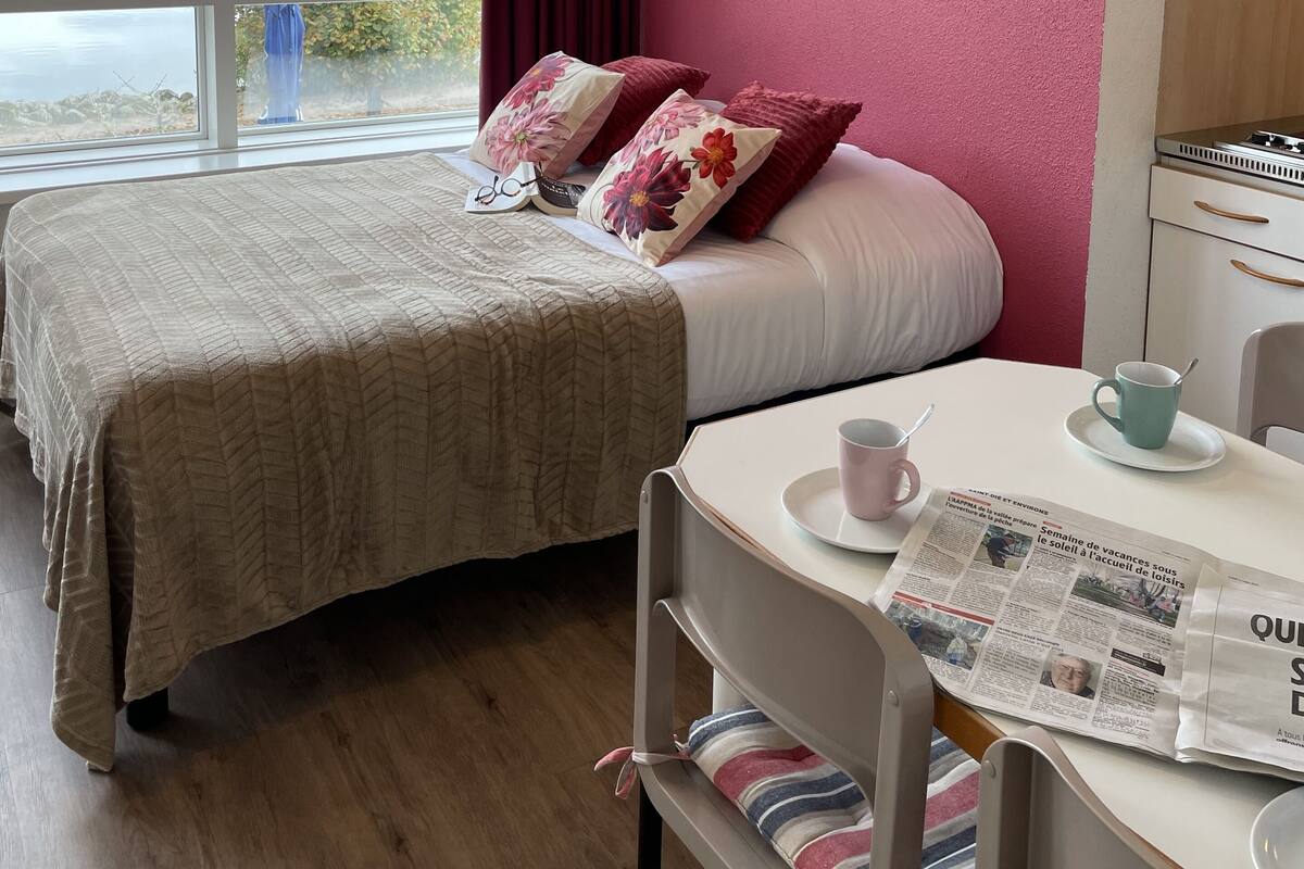 A hotel room with a bed, a table with a newspaper, and a view of a lake and mountains.