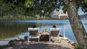 Dos personas sentadas a una mesa bajo una carpa, con vista a un lago y una ladera boscosa al fondo.