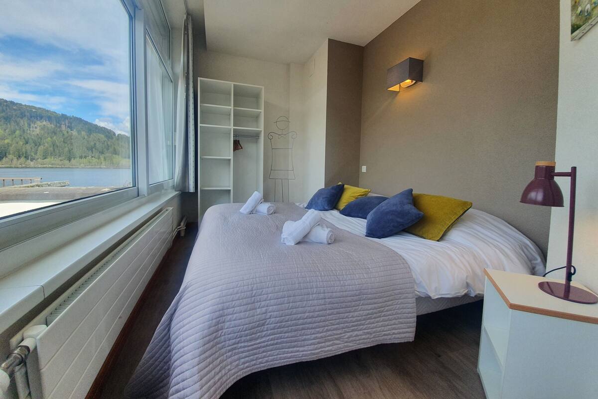 A bedroom with a large window, a bed with pillows, a bedside table, and a wardrobe.