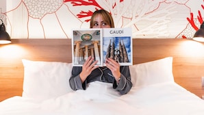 A person holding a book titled 'GAUDÍ' in a room with a wooden headboard and a wall with abstract red and white patterns. - ibis Styles Barcelona City Bogatell (Barcelona)